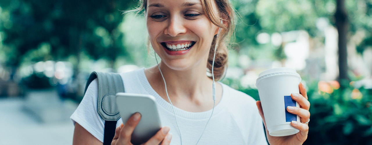 smiling young beautiful woman walking, holding drink, wearing headphones and having video call on smartphone on street