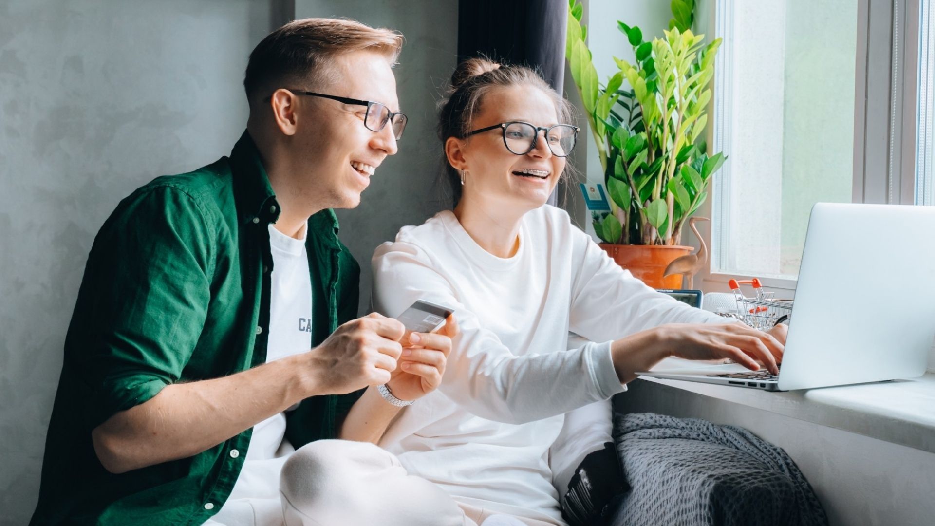 Happy couple holding credit card looking at laptop laughing relaxing sit on sofa making purchases