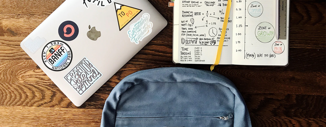 apple macbook with stickers on it and a binder and backpack on wood table